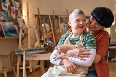 Portrait of smiling young woman embracing boy with disability while enjoying art class in studio copy space clipart