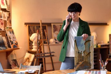 Kadın sanat uzmanının belden yukarı portresi telefonla konuşuyor ve galeride resim tutuyor.
