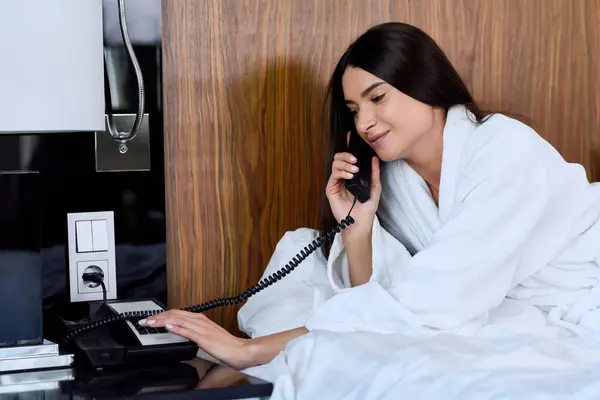 穿着浴衣的年轻女人躺在床上用手机在宾馆房间里微笑 免版税图库照片