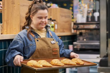 Waist up portrait of smiling young woman with Down syndrome enjoying work in cafe and holding tray with fresh pastry copy space