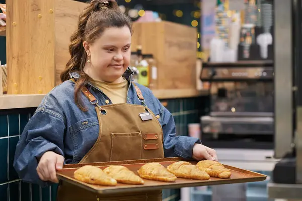 Waist up portrait of smiling young woman with Down syndrome enjoying work in cafe and holding tray with fresh pastry copy space