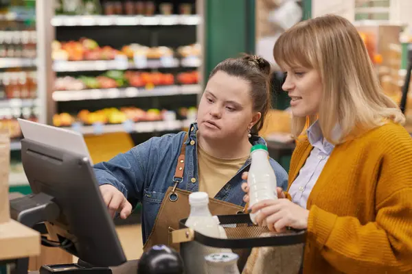 Waist up portrait of young woman with Down syndrome helping customer at self checkout while working in supermarket
