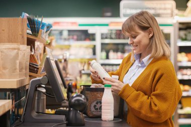 Side view portrait of smiling Caucasian woman using self checkout machine buying milk and groceries in supermarket 
