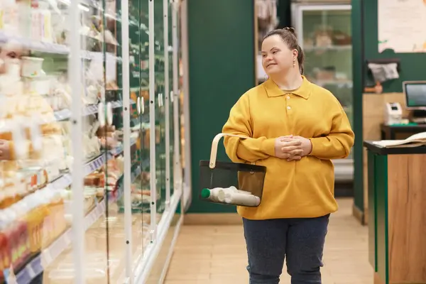 Portrait of smiling young woman with Down syndrome enjoying grocery shopping in supermarket and standing by freezer display copy space