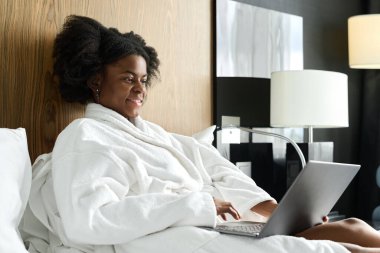 Genç Afro-Amerikan kadın portresi bornoz giyer ve otel odasında dizüstü bilgisayar kullanarak dinlenir.