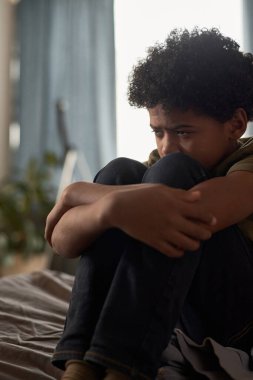 Ağlayan gencin dikey portresi. Odasında oturmuş dizlerine sarılıyor.