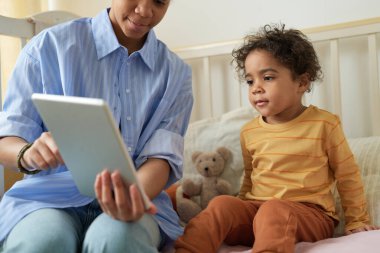 Afro-Amerikalı ebeveyn, çocuk dijital oyunlarını tablette, yatakta otururken ve etkileşimde bulunurken gösteriyor. Doldurulmuş oyuncaklarla dolu bir oda ve sıcak bir ortam.