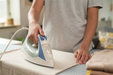 Ütü masasındaki giysileri ütüleyen kişi pencerenin yanındaki elektrikli ütüyle iyi ışıklandırılmış yastıklı, rahat oda ayarlı ve desenli yastıklı.