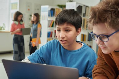 Okul kütüphanesinde arkadaşlarıyla dizüstü bilgisayar kullanan genç bir çocuğun portresi ve araştırma projesi.