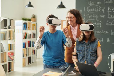 Modern okul sınıfında VR kullanan iki çocuğun belden yukarı portresi ve fotokopi alanında yardımcı bayan öğretmen.