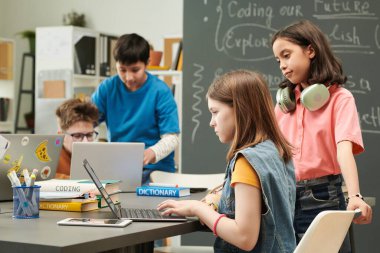 Okul kütüphanesinde bilgisayar kullanan ve ön planda fotokopi alanındaki iki kıza odaklanan çocukların renkli portresi
