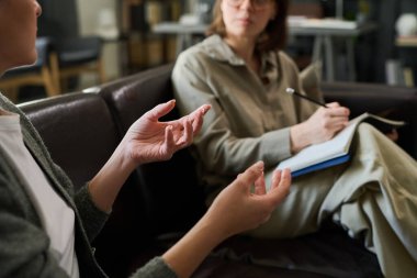 Two individuals engaging in counseling session on couch, one holding clipboard and pen, focusing on discussion, expressing thoughts with hand gestures clipart