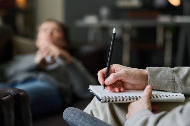 Hasta arka planda yüzü bulanık otururken terapist elinde kalem ve not defteri tutuyordu. Hastalara ayrıntıları göstermeden el ve kaleme odaklan