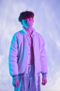 Mavi ve mor arkaplanlı bir stüdyoda beyaz ceket ve gümüş pantolon giyen genç bir adam portresi, ciddi bir ifadeyle kameraya bakıyor.