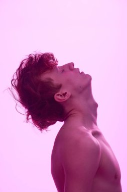 Kıvırcık saçlı genç adam başını yukarı doğru eğerek pembe arka planda siluet etkisi yaratıyor ve kaslı üst vücudunda yumuşak gölgeler oluşturuyor.