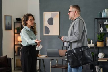 Üst düzey bir adam elinde bir cihaz tutarken genç bir bayanla iş birliği yapıyor. Ofis arka planında raflar, dekoratif eşyalar ve duvarda büyük bir tablo yer alıyor.