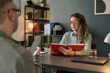 İki meslektaş ofis masasında karşılıklı otururken profesyonel tartışmalar yapıyorlar. Arka planda modern kitaplık ve klasörler var. Bir tanesi kırmızı klasör.