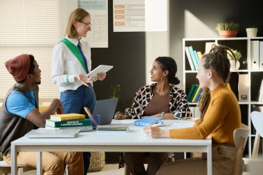 Üniversitede sınıfta masada oturan çeşitli öğrencilerle konuşan gülümseyen bir bayan öğretmenin yan görünümü.