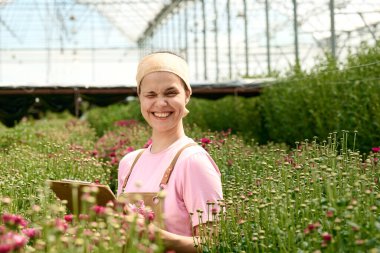 Çiçek plantasyonunda çalışırken ve elinde dosya kopyalama alanı tutarken gün ışığında göz kırpan sevimli genç bir kadının samimi portresi.