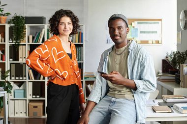 Ofis ortamında arka planda raflar ve bitkilerle işbirliği yapan gülümseyen meslektaşlar masasında cihazlar ve evrak işleriyle modern ve rahat bir atmosfer yaratıyorlar.