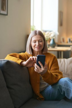 Modern olgun bir kadının dikey portresi akıllı telefon tutarken ve evdeki kanepede otururken ekranı dinlerken.
