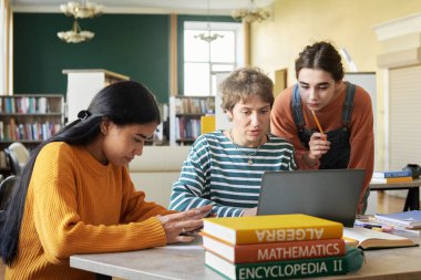 Bir grup öğrenci kütüphanede birlikte çalışıyor, dizüstü bilgisayarlar üzerinde çalışıyor ve notlar alıyorlar. Kitaplarla çevrili akademik bir ortamda çeşitli gruplar tartışıyorlar