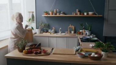 Orta yaşlı, beyaz bir kadının modern mutfakta içi doğal ışık, bitki ve taze ürünlerle dolu kahverengi kağıt torbalardan eşyalar düzenleyip sıcak ve sade bir atmosfer oluşturduğu bir fotoğraf.