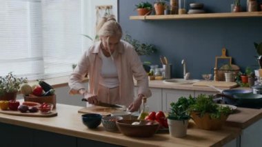 Orta yaşlı, beyaz bir kadının, ahşap tezgahta taze malzemelerle dolu sıcak bitki dolu bir mutfakta sağlıklı yemek pişirirken kaseye doğranmış soğan dökerken çekilmiş bir fotoğrafı.