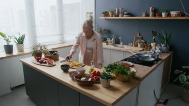 Orta boy beyaz kadın şefin, ahşap tezgah ve yeşil bitkilerle süslenmiş, rahat mutfakta taze sebzelerden sağlıklı yemekler yaptığı bir fotoğraf.