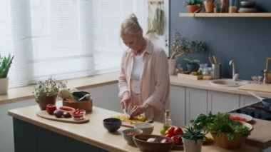 Yetişkin beyaz bir kadının ahşap tahtada sarı dolma biber keserken orta boy fotoğrafı. Modern, bitki dolu, penceresinden güneş ışığı gelen mutfakta duruyor.