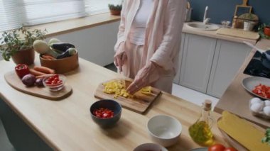 Çamaşır giysisi içinde tanınmayan bir kadının, parlak, rahat mutfak ortamında, doğal ışık ile süslenmiş, ahşap tahtada sarı biber doğrarken görüntüsü.