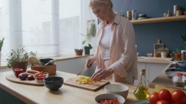Orta boy beyaz kadın, mutfak ortamında ahşap tezgahın yanında durup cam kaseye doğranmış sarı dolma biber dökerken, evde sağlıklı bir yemek hazırlarken.