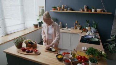 Orta yaşlı, beyaz bir kadının modern mutfağın ahşap tezgahında patlıcan keserken çekilmiş yeşil bitkiler ve doğal ışıkla süslenmiş bir fotoğrafı.