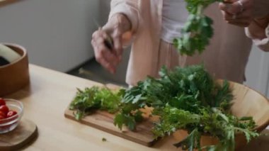 Gündüz vakti modern mutfak ortamında taze yeşil otları ahşap kesme tahtasıyla doğrayan tanınamayan kadın şefin yakın çekimi.