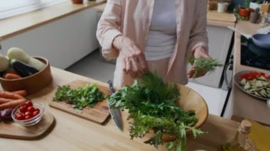 Ahşap tezgahın üzerinde maydanoz ve dereotları doğrayan, tabaklar ve kâselerdeki taze malzemelerle dolu, tanınamayan kadın aşçı.