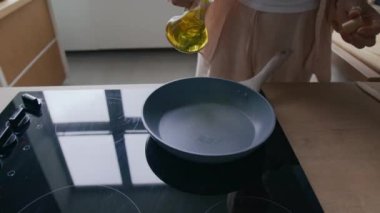 Tanımlanamayan bir kadının, ahşap tezgahlar ve işlevsel pişirme alanlarıyla modern mutfaktaki tavaya zeytinyağı koyması.