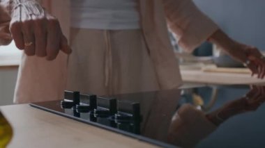 Tanımlanamayan yetişkin kadın elinin modern sobanın düğmesini ayarlaması ve ısıtma için tavayı yerleştirmesi, minimalist mutfak ortamında konforlu ve işlevsel alanla yemek pişirmesi.