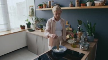 Yetişkin beyaz kadın aşçının sıcak mutfak ortamında, pencerelerden ve ahşap tezgahlardan gelen parlak ışıkla kızartma tavasında soğan pişirirken orta boy pozu.