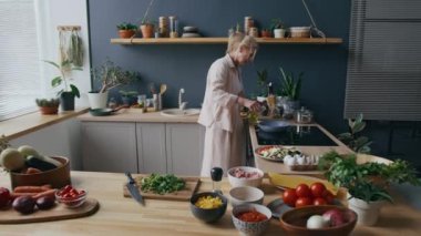 Kafkasyalı yetişkin bir kadının modern sobanın başında, ahşap tezgahın üzerinde taze malzemelerle, sıcak tavaya yağ eklerken orta ölçekli bir fotoğrafı.