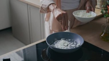 Modern indüksiyon sobasında dilimlenmiş soğanlı kızartma tavası cızırdıyor. Yakınlarda tanınmayan kadın aşçı doğal ışık ve ahşap tezgahlı minimalist mutfakta malzemeler hazırlıyor.