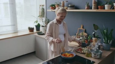 Orta yaşlı, beyaz bir kadının dolgun sebzeler pişirdiği, dolmalık biberler, domatesler ve havuçlar dahil, modern fırının tepesinde, parlak, rahat bir mutfakta.