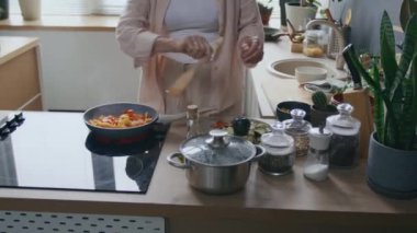 Tanımlanamayan kadın şefin modern sobada tavada cızırdayan sebzelere tuz eklemesi, sıcak mutfakta sağlıklı yemek pişirmesi.