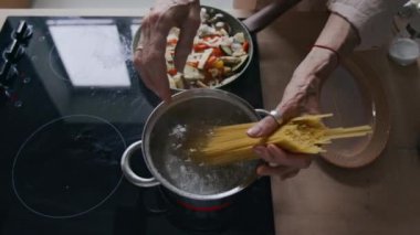 Tanınmayan kadın şefin, spagettiyi kaynayan suya atarken sebzelerin tavada cızırdadığı modern mutfakta ahşap tezgah ve soba tezgahıyla yemek pişirdiği tepeden aşağı görüntü.
