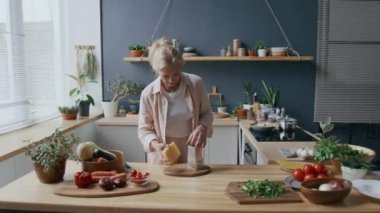 Orta yaşlı, beyaz bir kadının ahşap mutfak tezgahının yanında durup ahşap tahtanın üzerinde peynir rendelerken rahat, evcil atmosferde akşam yemeği hazırlarken görüntüsü.