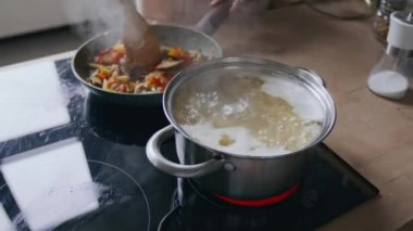 Tanınmayan birinin tavada sebze kızartması, ahşap kaşıkla karıştırması, modern mutfağın tepesinde kaynayan spagetti, parlak, rahat mutfak ortamında.