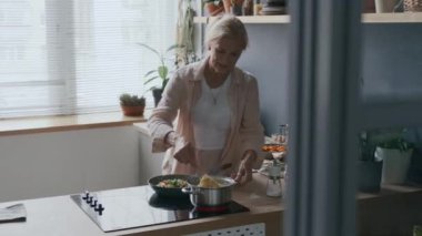 Orta yaşlı, beyaz bir kadının spagettiyi kaynayan suda karıştırdığı ve sonraki ocakta sebze kızarttığı doğal ışık dolu sıcak mutfakta akşam yemeği hazırlama sürecinin keyfini çıkardığı bir fotoğraf.