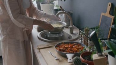 Modern mutfakta sebze ve mutfak gereçleri ahşap tezgahta dururken spagetti bolonez yaparken tanınmayan bir kadının pişirilmiş makarnayı durulaması.