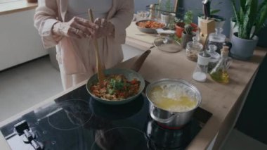 Tanımlanamayan bir kadının modern fırında tavada kızartılan sebzelere doğranmış otlar ve yakınlarda kaynayan spagetti eklenmesi.
