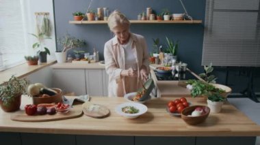 Orta boy beyaz kadın, rahat keten kıyafetler giyip, beyaz tabakta iki zarif tabak servis ederken, bitki dolu mutfakta, doğal ışıkla süslenmiş.