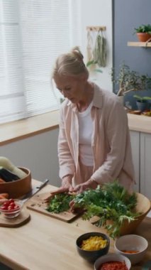 Kafkasyalı orta yaşlı bir kadının rahat bir şekilde günlük kıyafetlerini giyip kesim tahtasına otlar yerleştirirken, parlak, rahat bir mutfakta sağlıklı yemek yaparken, bitki ve çatal bıçaklarla süslenmiş, dikey bir fotoğraf.